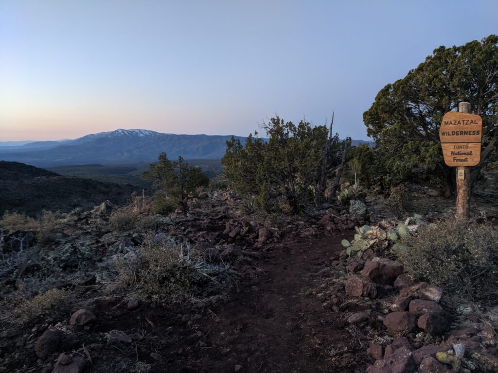 Twilight descends over the rugged terrain of the Mazatzal Wilderness, with a clear large wooden sign marking the entrance to the Tonto National Forest. The landscape is a rich tapestry of desert flora, including cacti and shrubs, with a mountain range in the background capped by lingering snow. The early morning light gently illuminates the scene, highlighting the natural beauty and serene atmosphere of this remote wilderness area.
