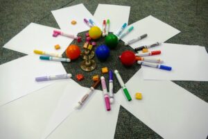A creative array of art supplies spread out on a carpeted floor. The scene includes several sheets of blank white paper arranged in a starburst pattern, surrounded by colorful markers scattered haphazardly. In the center, a Hanukkah dreidel is flanked by four small, bright rubber balls in orange, green, blue, and red. Scattered among the markers are star-shaped erasers in vivid yellow, adding a playful touch to the composition.