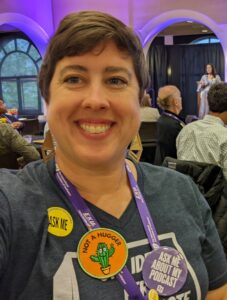 The author smiling at an IAF conference, wearing a 'Not a Hugger' badge with a cactus illustration, a 'Ask Me About My Podcast' button, and an 'Ask Me' sticker. They have short hair, geometric earrings, and a lanyard around their neck. There are people seated in the background with a speaker on stage.