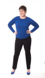 Sharon Tewksbury-bloom standing with hands on hips wearing a bold blue sweater, black pants and blue heels. Smiling and engaging.